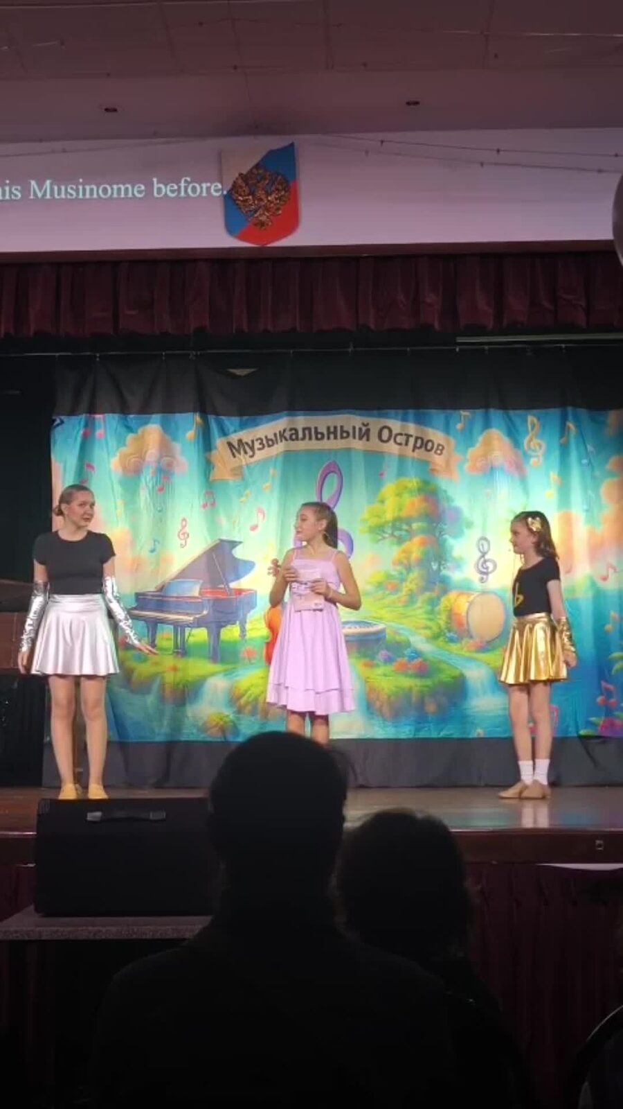Three students performing together on stage in front of the Musical Island Productions backdrop